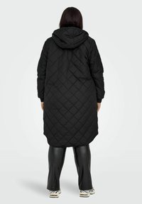 Schwarzer, gesteppter langer Mantel mit Kapuze, mit einem Rautenmuster, seitlichen Taschen und abgerundetem Saum. Getragen über schwarzen Leggings und Schuhen.