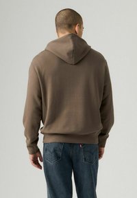 Sudadera con capucha marrón de corte relajado, con puños y dobladillo acanalados que caen sobre unos jeans de mezclilla oscuros. El diseño presenta un sencillo detalle en la parte posterior.