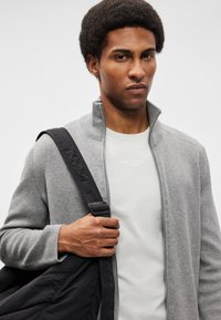 Γκρι πλεκτό πουλόβερ με φερμουάρ και ψηλό γιακά, επικαλυμμένο πάνω από ένα ανοιχτόχρωμο μπλουζάκι. Σκούρα τσάντα ώμου με ιμάντα ακουμπάει στο χέρι.