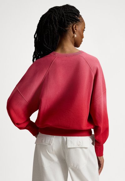 Overdimensjonert genser med et gradientmønster fra rødt til lysere rosa. Laget av mykt materiale med ribbede mansjetter og nederkant. Kombinert med hvite bukser.