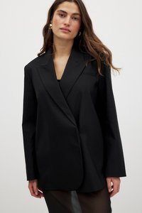 Mustat bleiseri, joka on valmistettu sileästä kankaasta, jossa on lovettu kaulus, pitkät hihat ja yksi nappi. Hieman ylipitkä, minimaalisen yksinkertainen.