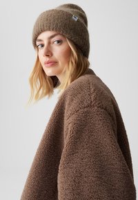 Braune Mütze mit einem gerippten Muster und einem Logo-Patch, kombiniert mit einer kuscheligen braunen Fleecejacke mit einer weichen, strukturierten Oberfläche.