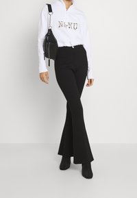 Sweat à fermeture éclair blanc avec logo "N>K>D" imprimé léopard et pantalon noir évasé. Le mannequin porte un petit sac noir en bandoulière et des bottes noires.