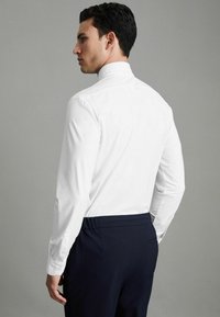 Vit långärmad skjorta med hög krage, med knäppning vid ärmsluten. Bärs med marinblå byxor som har en elegant, åtsittande design.
