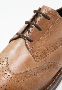 Chaussure brogue en cuir marron avec lacets noirs, perforations détaillées et couture en zigzag sur l'avant et les côtés.