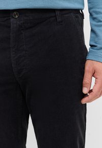 Czarne spodnie z korduroju mają żeberkową fakturę, dwie przednie kieszenie, zapięcie na guzik oraz dopasowany krój, częściowo pokazane obok ręki.