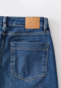 Jean en denim bleu avec une patch en cuir à l'arrière comportant un logo. Le tissu est texturé avec un léger éclat, et les coutures sont contrastées.
