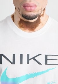 Baltos medvilnės marškinėliai su juodu 'NIKE' ženklu ir mėlyna swoosh dizainu. Užfiksuota artima apžvalga, rodomi kaklo ir smakro odos tekstūros taškeliai.