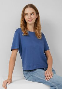 Jeune femme aux cheveux châtains clairs portant un t-shirt bleu à manches courtes et un jean bleu clair, assise sur un bloc blanc, souriant et regardant à droite.