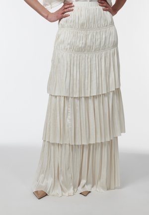 Persona con una falda larga, de varias capas, plisada y de color crema, con las manos en las caderas; se ven unos zapatos beige de punta en un fondo blanco liso.