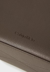Sac en cuir brun texturé avec le logo "Calvin Klein" en relief. Présente des lignes épurées et des détails de couture subtils.