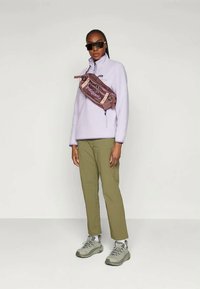 Ljuslila fleecetröja med halvdragkedja, olivgröna byxor, gråa klumpiga sneakers och brun axelremsväska med logotypdetalj.