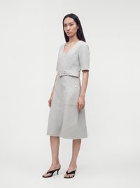Femme portant une robe midi gris clair ceinturée, avec des manches aux coudes et des sandales noires à talons, debout devant un fond blanc uni.