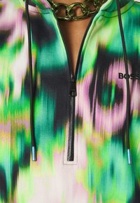 Sweat à capuche tie-dye coloré vert, violet et noir avec fermeture éclair noire et cordons, partiellement ouvert, associé à un gros collier chaîne dorée.