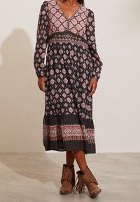 Kvinna iklädd en långärmad svart midiklänning med rosa och vita geometriska mönster samt bruna fransade ankelstövlar, stående inomhus.