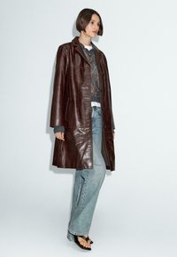 Cappotto lungo in pelle marrone con una texture leggermente lucida, abbinato a un maglione grigio oversize e jeans azzurri chiari. Apertura frontale, chiusura con un singolo bottone.