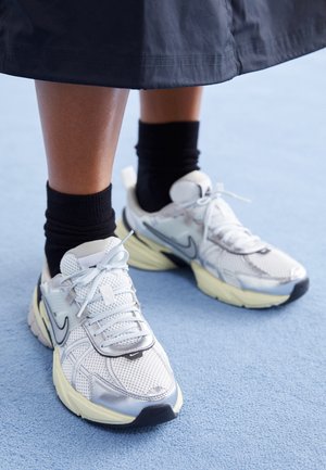 Osoba nosząca czarne skarpetki, białe i srebrne buty sportowe Nike, stojąca na jasnoniebieskim wykładzinowym podłodze z widoczną częścią ciemnej spódnicy.