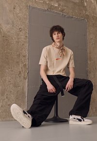 Beige katoen T-shirt met oranje logo, zwarte baggy broek en witte sneakers met marineblauwe accenten. Gestyled met een patroon sjaal.