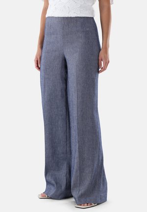 Frau trägt hoch taillierte, weit geschnittene blaue Leinenhosen kombiniert mit einem weißen Spitzenoberteil und offenen Sandalen, steht vor einem schlichten Hintergrund.