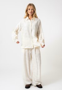 Camicia bianca, testurizzata, con bottoni, spalle basse e vestibilità rilassata, abbinata a pantaloni bianchi larghi e scarpe marroni.