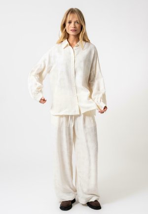 Chemise blanche texturée à boutons avec des épaules Tombées et une coupe décontractée, associée à un pantalon blanc ample et des chaussures brunes.