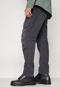 Svarta cargo-byxor med en avslappnad passform, som har flera fickor, texturerat tyg och synliga sömdetaljer. Bärs med svarta stövlar.