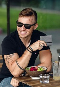 Šedá peněženka, kterou drží muž v černém tričku, s brýlemi na očích a tetovanou paží, sedící u stolu se salátovou miskou.