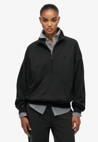 Czarna bluza z zamkiem 1/4 o oversize'owym kroju, miękkiej teksturze, z kieszenią z przodu i dyskretnym logo. Połączona z koszulą z kołnierzykiem w paski.