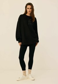 Czarna, oversize bluza z wytłoczonym napisem, zestawiona z czarnymi legginsami, kremowymi sneakersami i beżowymi skarpetkami z napisem. Gładka tekstura.