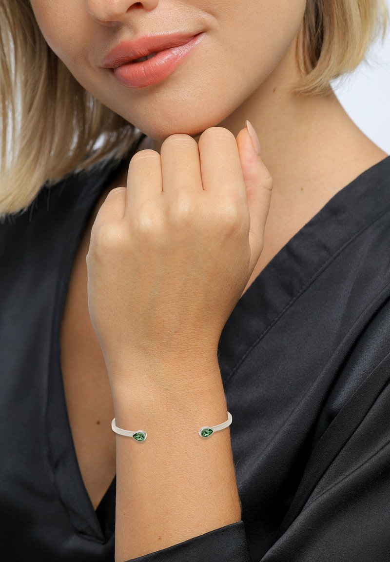 Silberne Armreifen mit grünen Edelsteinen an jedem Ende, vor einem hellen Hautton und einem schwarzen Stoffhintergrund präsentiert.