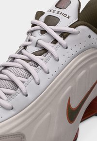 Gros plan d'une basket blanche Nike Shox montrant les lacets, la languette, le côté perforé et le logo Swoosh de Nike en orange et vert foncé.