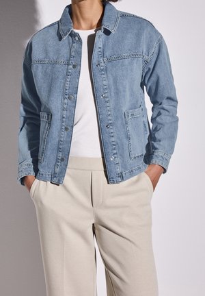 Person iført lyseblå denimjakke over hvid top og beige bukser med hænderne i lommerne mod en ensfarvet baggrund.