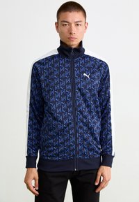 Marineblå zip-up jakke med gentagne blå logoer, hvide sidelister, høj krave og ribbede manchetter og kant. Blødt, glat materiale.