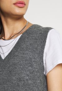Gilet grigio a maglia con scollo a V sopra una t-shirt bianca. Tre sottili collane d'argento sono sovrapposte attorno al collo. Tessuto morbido, design casual.
