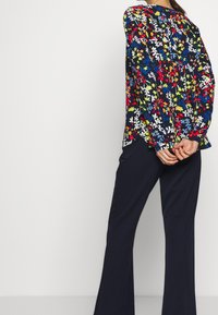 Kwiatowa bluzka z długim rękawem, na ciemnonawowym tle, z żywymi czerwonymi, żółtymi i niebieskimi kwiatami. Połączona z czarnymi spodniami o szerokim kroju.