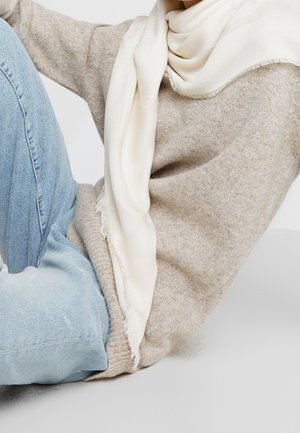 Personne portant un pull beige, un jean bleu clair et une écharpe crème, assise les jambes pliées contre un fond clair uni.