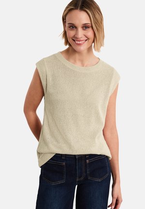 Femme souriante aux cheveux blonds courts, portant un haut en maille beige sans manches et un jean bleu foncé, debout les mains derrière le dos.