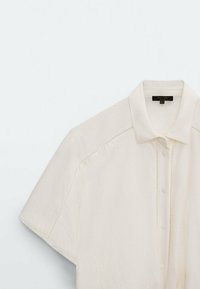 Kurzärmliges, cremefarbenes Hemd mit Knopfleiste und subtilen strukturierten Details an den Schultern und der Knopfleiste.