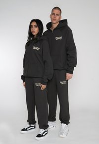 Graue Kapuzenpullover und -hosen mit einem Logo auf der Brust und dem Bein, vor einem neutralen Hintergrund. Sneakers runden den Look ab.