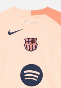 Helles Pfirsichfarbenes Nike-Trikot mit dem dunkelblauen FC Barcelona-Wappen, einem großen schwarzen Spotify-Logo und orangefarbenen Schulterakzenten.