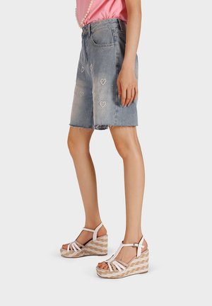 Gonna di jeans con orlo sfrangiato e decorazioni a forma di cuore, abbinata a sandali con zeppa bianchi e fibbie, caratterizzati da una piattaforma testurizzata.