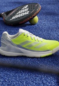 Bright yellow and white Adidas sports shoe on blue textured court surface next to a black paddle racket and a yellow ball.