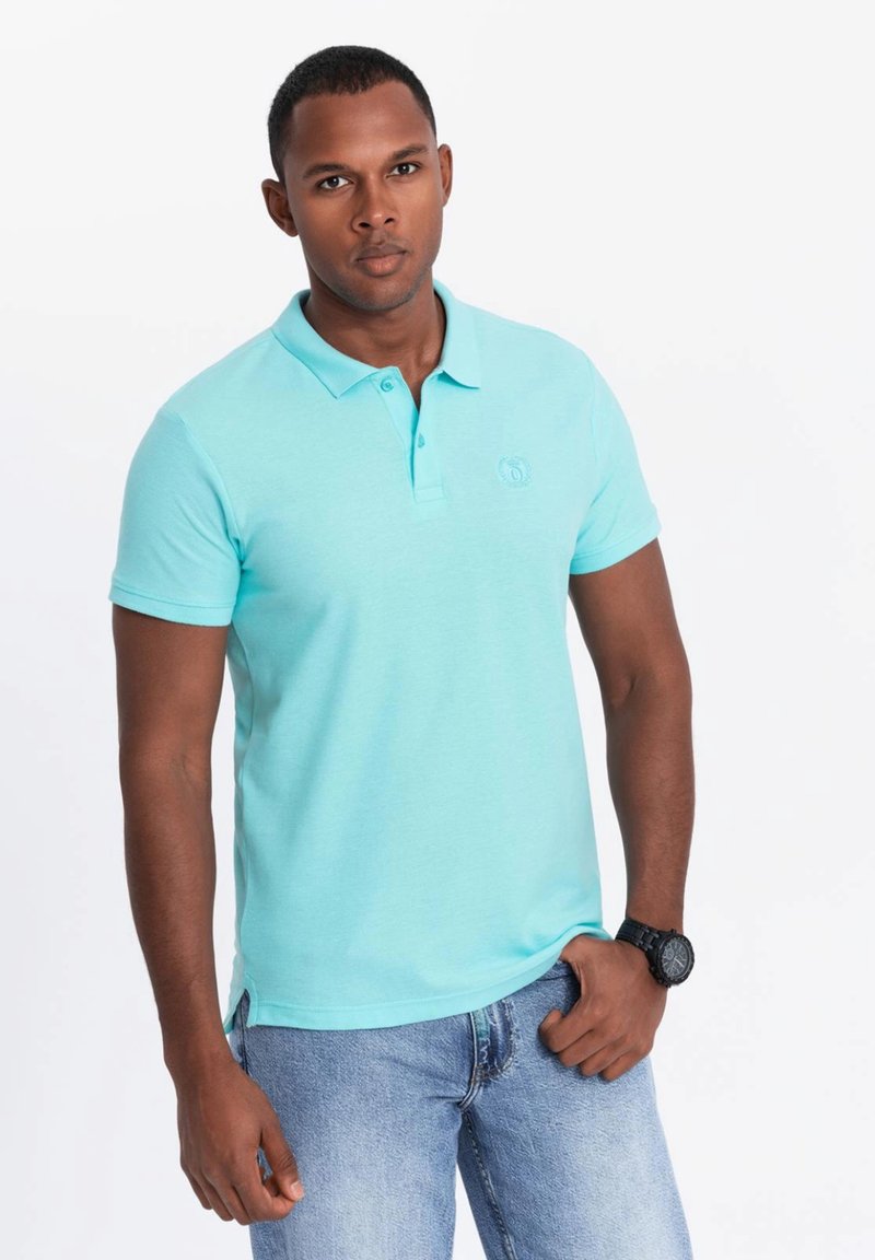 Světle modré polo tričko z bavlněné směsi, s dvoupoložkovým zapínáním a jemným logem, v kombinaci s denimovými džíny a černými hodinkami.