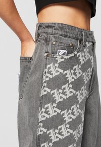 Grå denimbukser med et gjentatt hvitt logomønster, med metallknapper, sidelommer og en lapp på baklommen.