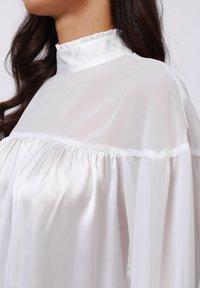 Long-sleeved blouse in sheer white fabric, featuring a ruffled high collar and gathered pleats across the chest for a soft silhouette.