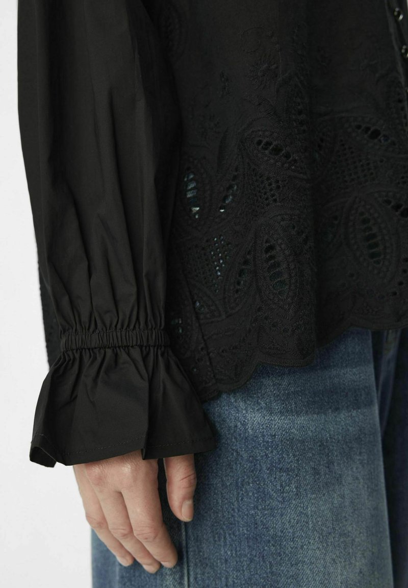 Haut noir avec broderie texturée arborant des motifs paisley, manches élastiques et design de manches évasées, associé à un jean en denim.