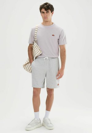 Junger Mann trägt hellviolettes T-Shirt, graue Shorts mit Kordelzug, weiße Socken, Sneakers und eine beigefarbene gestreifte Tragetasche über der Schulter.