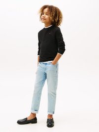 Black long-sleeve shirt with white trim, light blue denim jeans rolled at cuffs, and black loafers. Curly hair, standing pose.