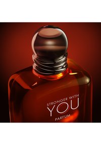 Bottiglia di profumo in vetro ambra con tappo nero rotondo, etichettata "Stronger With You Parfum" su uno sfondo rosso scuro.