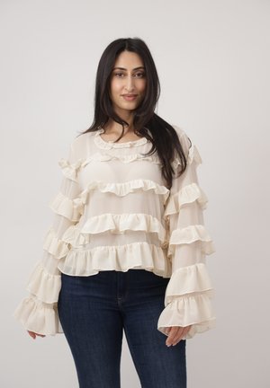 Femme aux longs cheveux foncés portant un chemisier à volants de couleur crème et un jean bleu foncé, debout devant un fond clair uni.